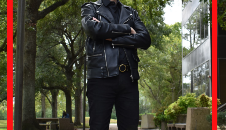 Carlo Mazziotta, Undergrad in Sociology, standing outside the Patio Café. The trees curving above him while light peaks through the trees. Dressed in an all black ensemble of a button up, slacks, and leather boots and a jacket.