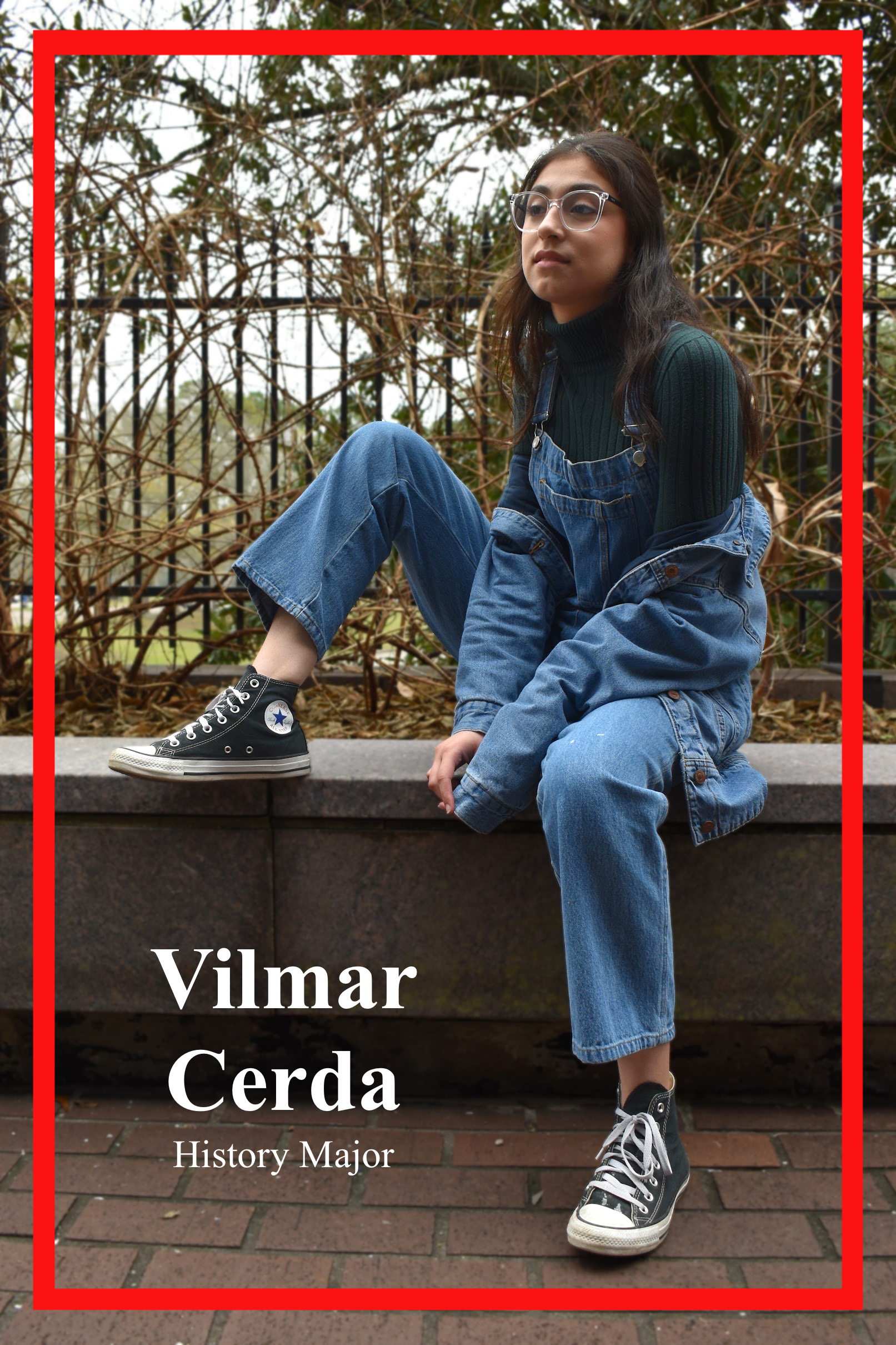 Vilmar Cerda, History Major, posed on a ledge dressed in all denim overalls and jacket, paired with a forest green turtleneck and black converse.
