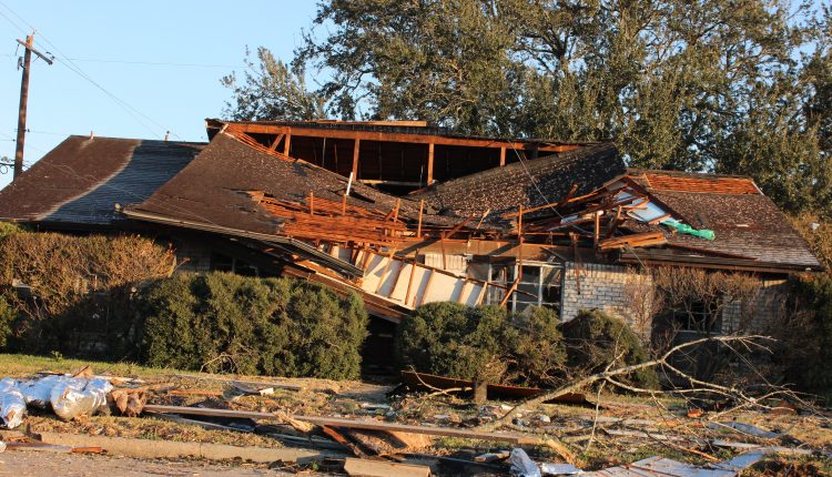 A house with the roof bowing in and partially ripped off surrounded by Debris.