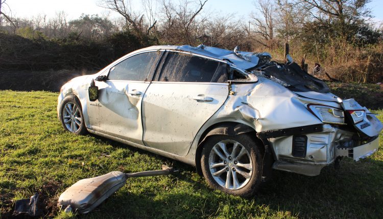 A silver car that is crushed in a field with the gas tank laying to the side of it.