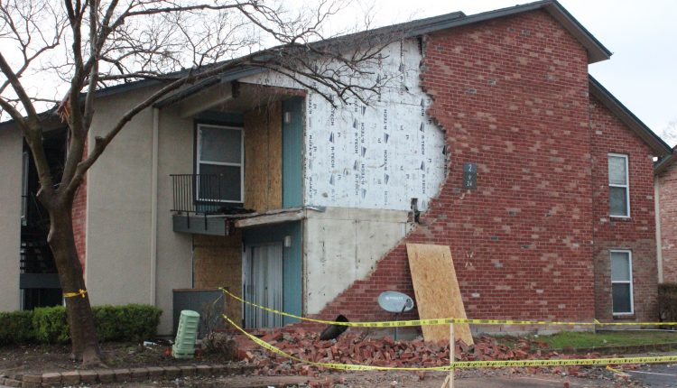 An apartment building in Deer Park missing brick siding with a pile of bricks below and caution tape around it. The balcony is barely hanging on the second floor.